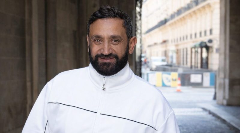Portrait d’un animateur de télévision français en veste blanche, regard caméra, debout dans une rue urbaine avec immeubles en arrière-plan, illustrant une révélation médiatique sur un contrat financier record dans le paysage audiovisuel