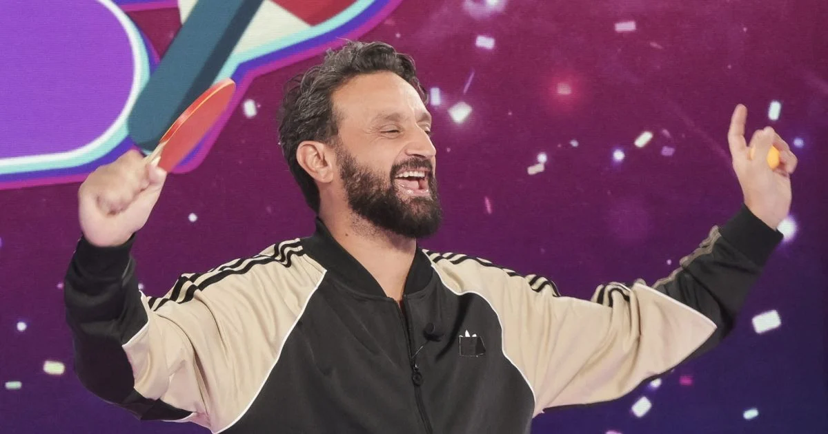 Animateur de télévision en plein plateau d’émission, souriant et levant les bras devant un décor lumineux et festif, illustrant le succès et l’audience d’un programme populaire du paysage audiovisuel français