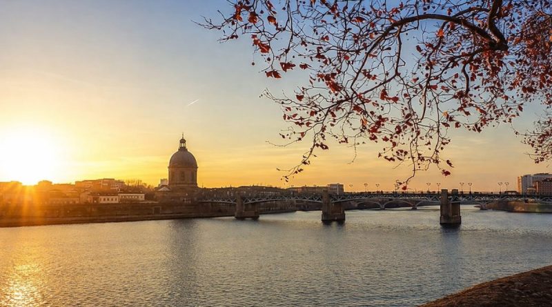 Grande rivière traversant une ville au coucher du soleil, avec un pont métallique au centre, un dôme ancien sur la rive et des branches d’arbre aux feuilles rouges au premier plan au-dessus de l’eau calme.