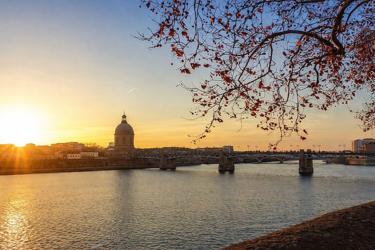 Grande rivière traversant une ville au coucher du soleil, avec un pont métallique au centre, un dôme ancien sur la rive et des branches d’arbre aux feuilles rouges au premier plan au-dessus de l’eau calme.