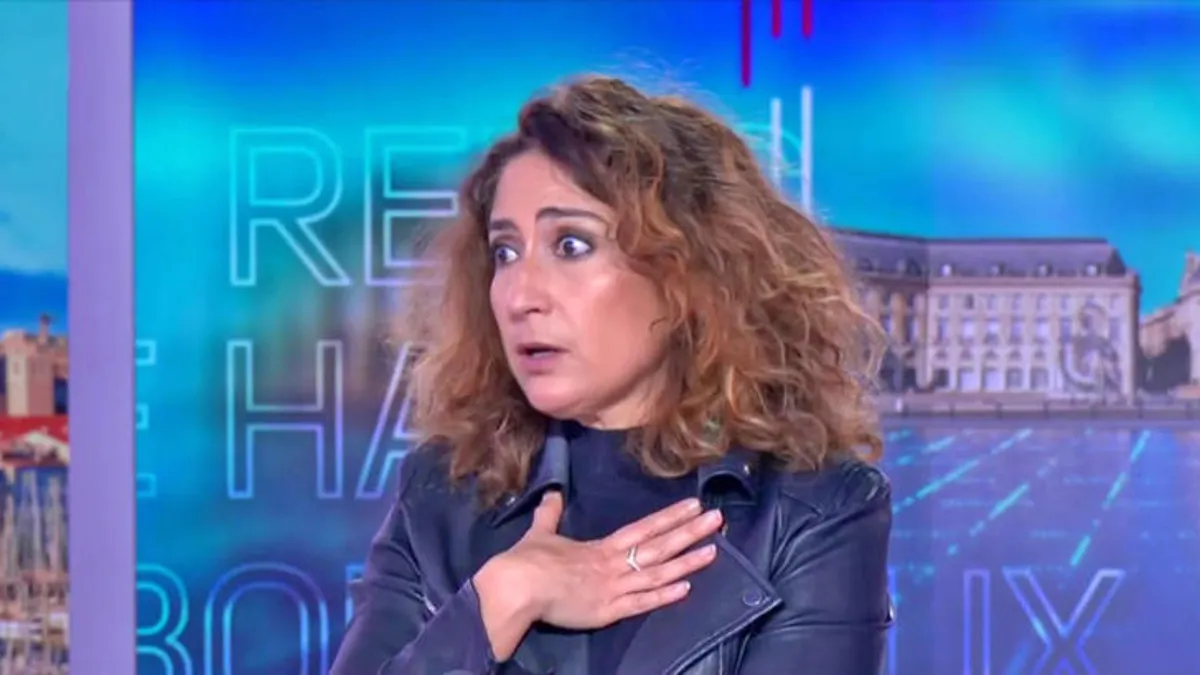 femme intervenante sur un plateau de télévision, regard surpris et main posée sur la poitrine lors d’un échange tendu en direct
