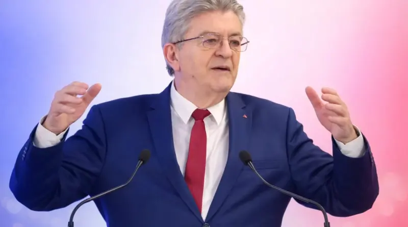 homme politique français aux cheveux gris s’exprimant derrière un pupitre avec deux micros, levant les mains lors d’un discours public sur une scène aux couleurs bleu, blanc et rouge