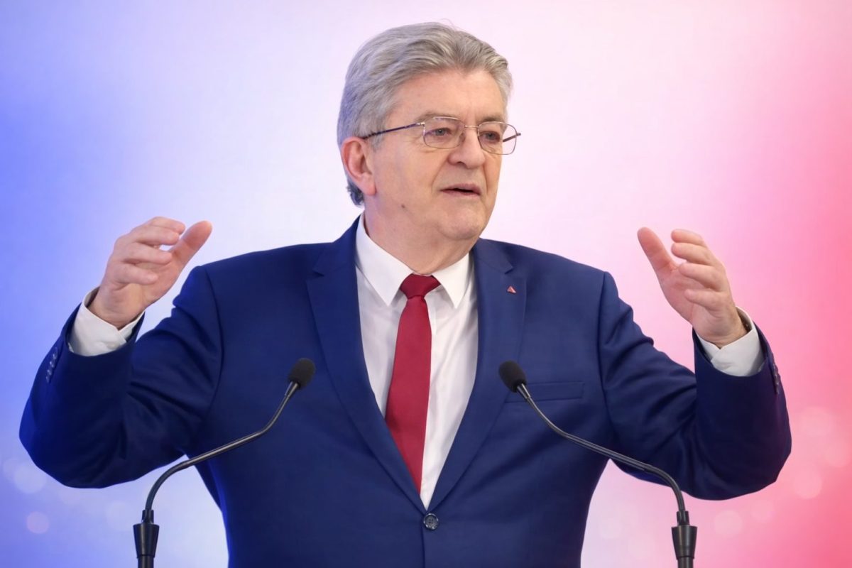homme politique français aux cheveux gris s’exprimant derrière un pupitre avec deux micros, levant les mains lors d’un discours public sur une scène aux couleurs bleu, blanc et rouge