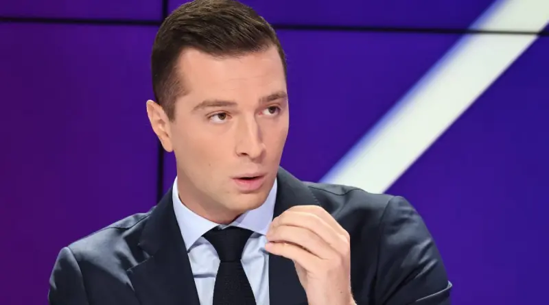 Un responsable politique en costume sombre s’exprimant en plateau télévisé, geste précis de la main, regard concentré, dans une ambiance de débat face à des critiques publiques