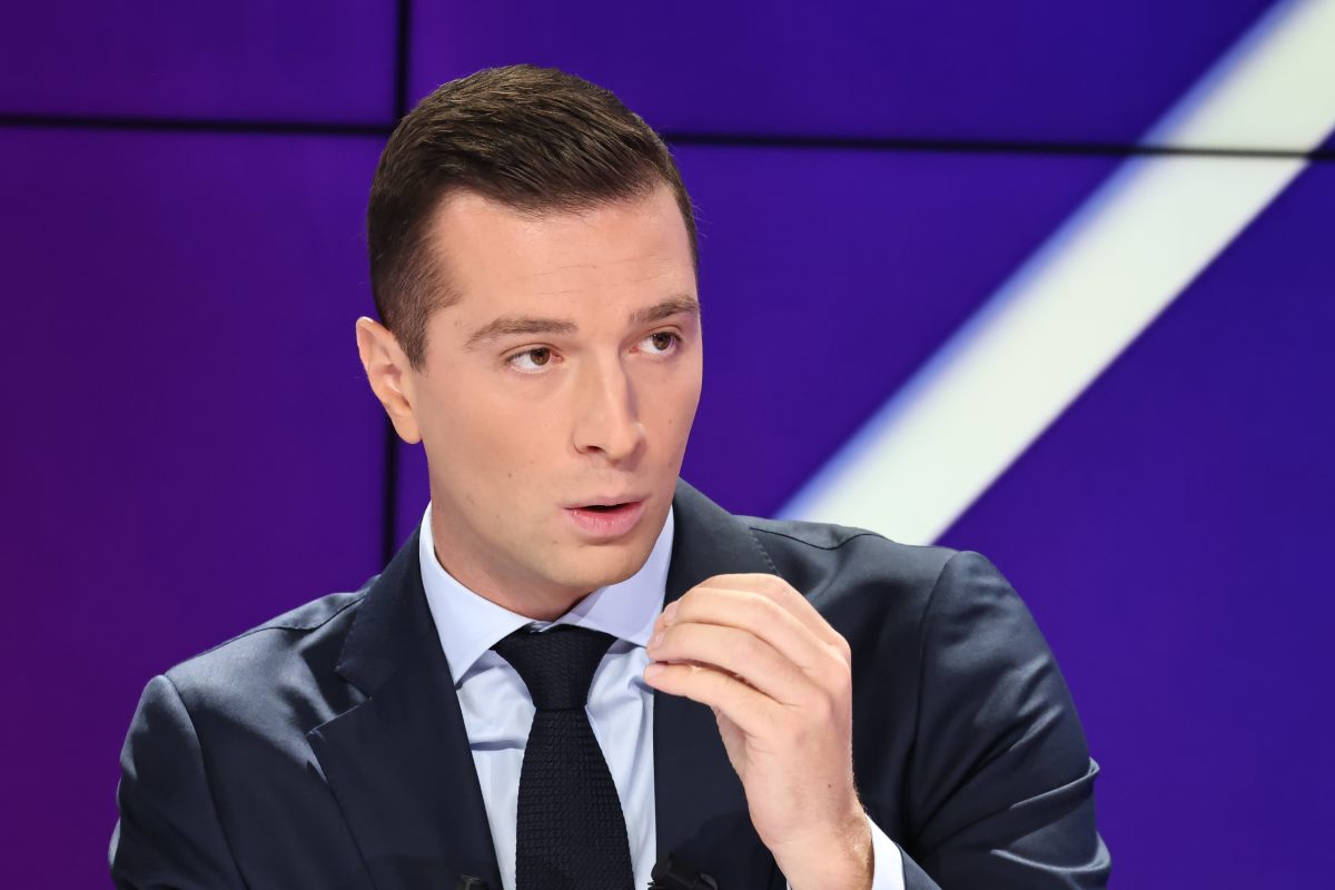 Un responsable politique en costume sombre s’exprimant en plateau télévisé, geste précis de la main, regard concentré, dans une ambiance de débat face à des critiques publiques
