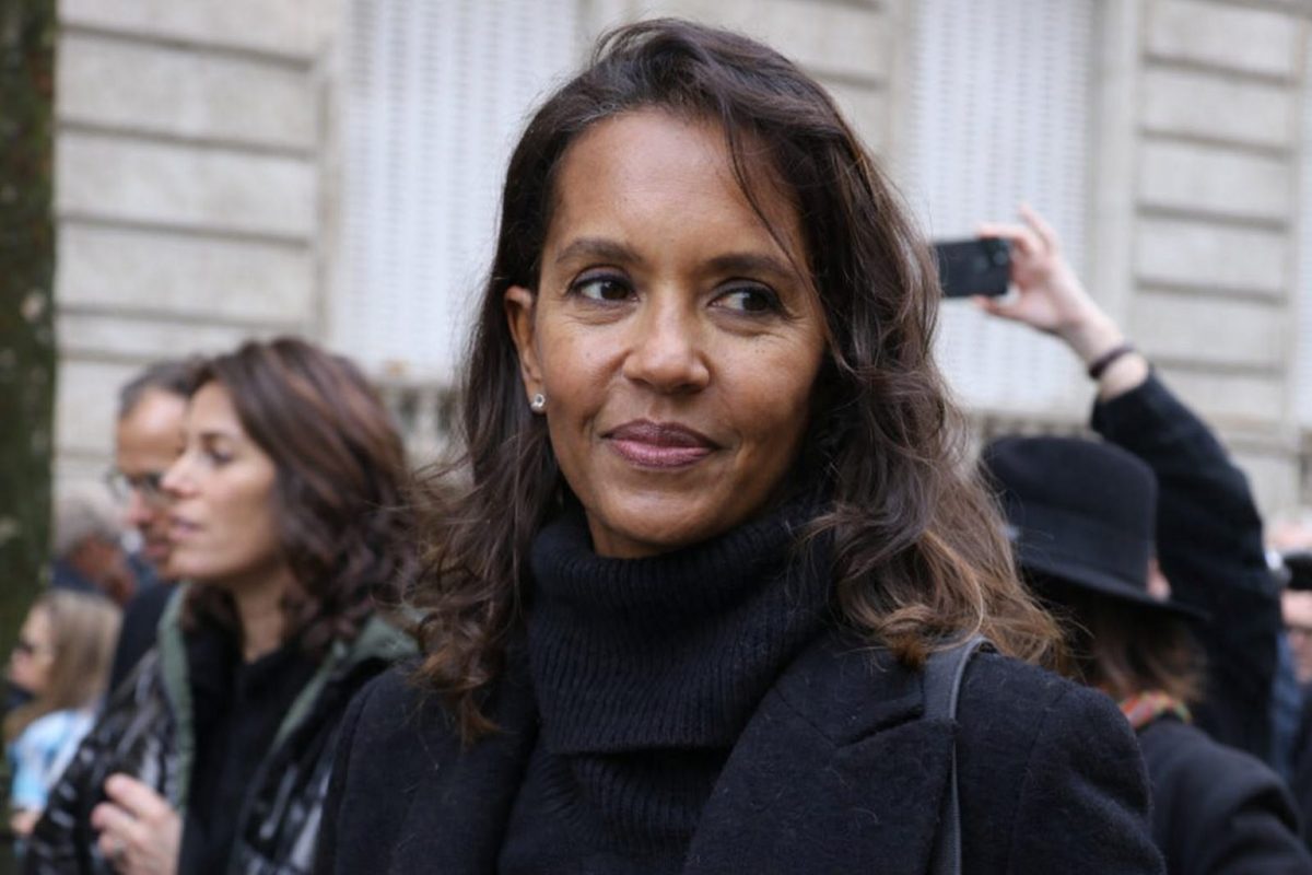 animatrice française en extérieur entourée de personnes regardant sur le côté avec une expression attentive, manteau noir, décor urbain flou en arrière-plan