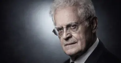 Portrait d’un homme politique âgé aux cheveux blancs, portant des lunettes et un costume sombre, regard sérieux et posé sur fond neutre