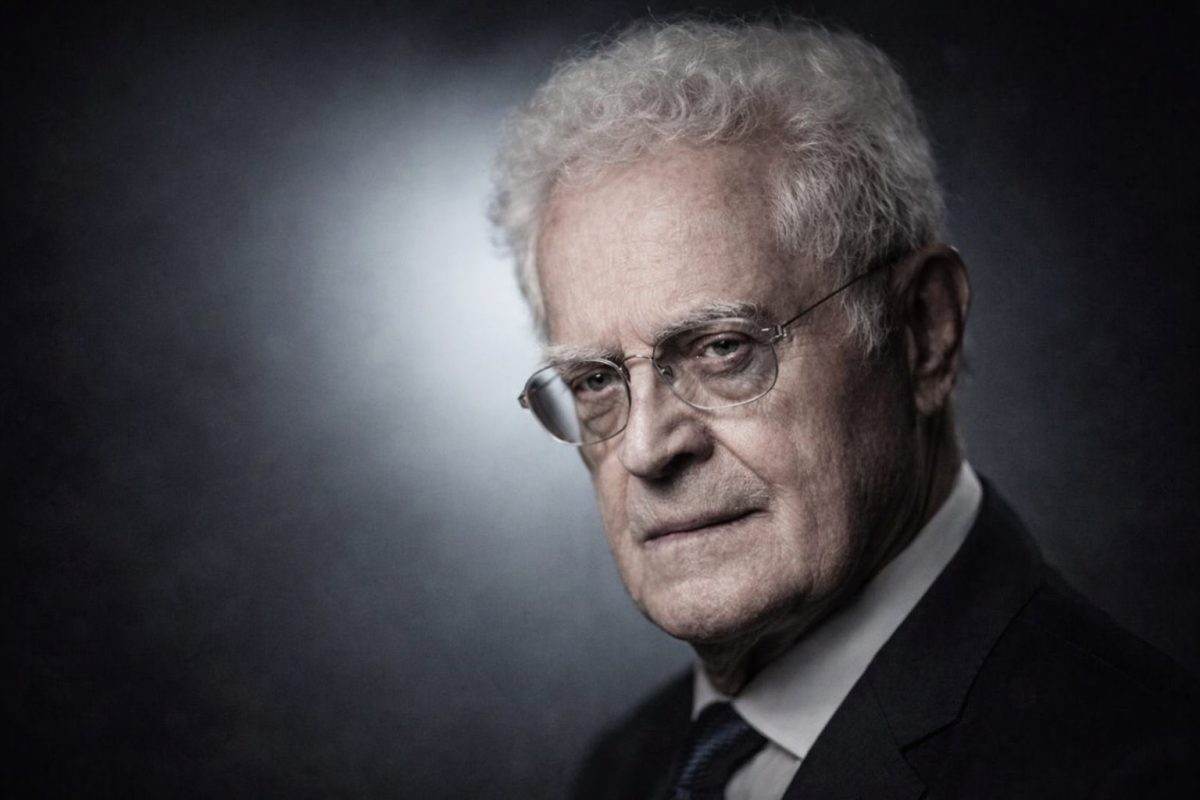 Portrait d’un homme politique âgé aux cheveux blancs, portant des lunettes et un costume sombre, regard sérieux et posé sur fond neutre