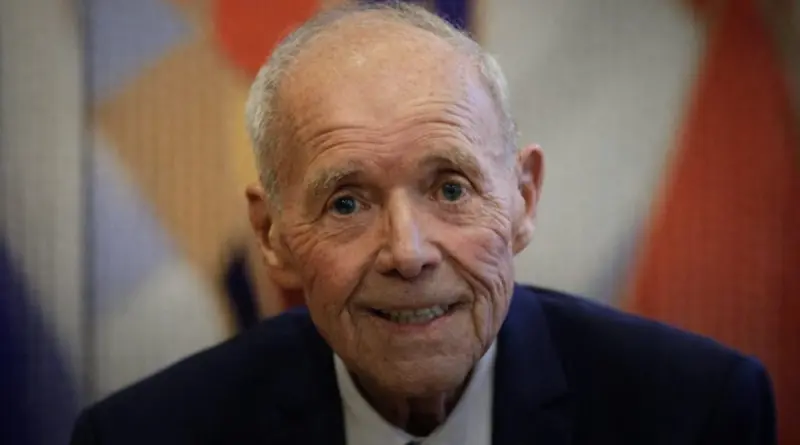 portrait d’un homme politique âgé souriant portant un costume sombre et une cravate, dans une salle officielle avec un arrière-plan flou aux couleurs républicaines, illustrant une réélection municipale