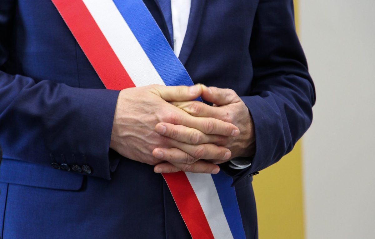 Gros plan sur un homme en costume portant une écharpe tricolore, mains jointes devant lui, symbole de fonction officielle et de responsabilité politique dans un contexte institutionnel