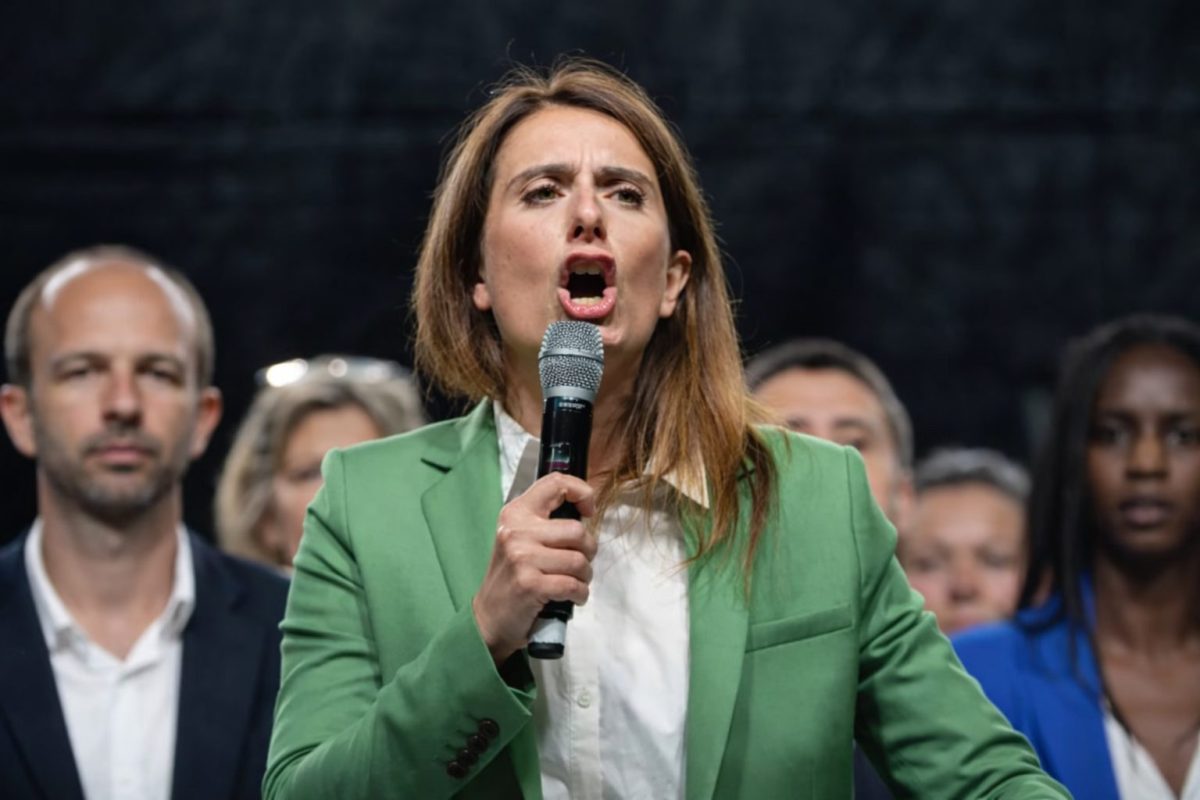 femme politique s’exprimant avec intensité au micro lors d’un meeting électoral entourée de plusieurs personnes en arrière-plan dans une ambiance de campagne municipale