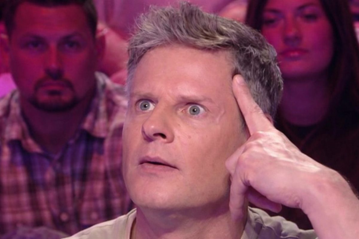 animateur de télévision français en plein débat sur un plateau télé, regard intense et doigt posé sur la tempe, avec un public assis en arrière-plan sous un éclairage rose et violet typique d’un studio de talk-show