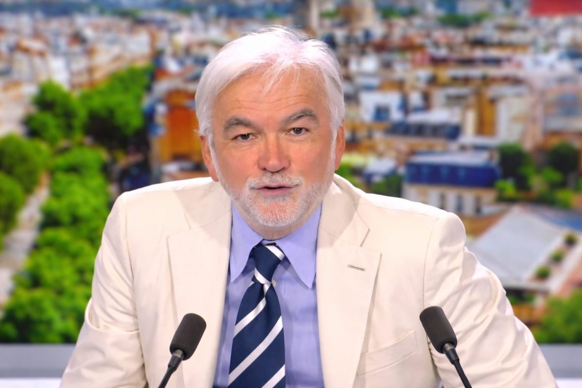 Présentateur de télévision aux cheveux blancs assis derrière un bureau sur un plateau d’émission d’actualité, devant un décor urbain flouté, participant à un débat télévisé avec deux microphones visibles au premier plan.