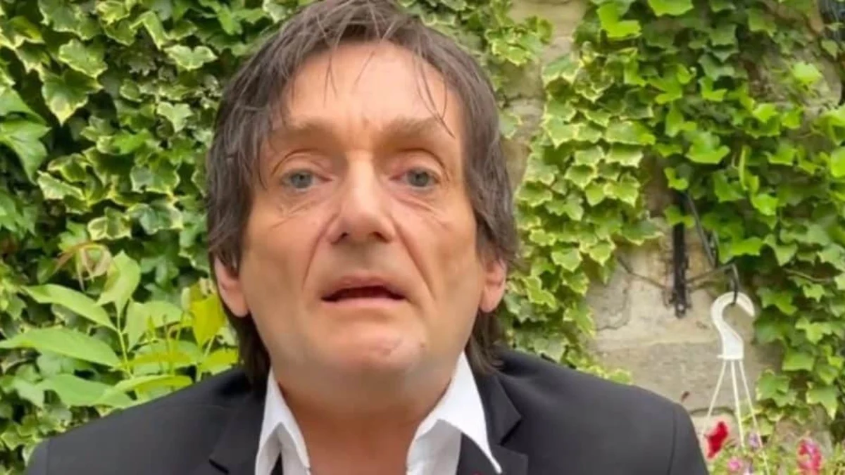 Un homme d’âge mûr au visage marqué et au regard fatigué s’exprime face à une caméra dans un jardin, devant un mur recouvert de plantes grimpantes vertes. Il porte une veste sombre et une chemise claire légèrement ouverte au col. Son expression paraît grave et préoccupée, comme s’il évoquait une situation personnelle difficile ou un moment de crise.