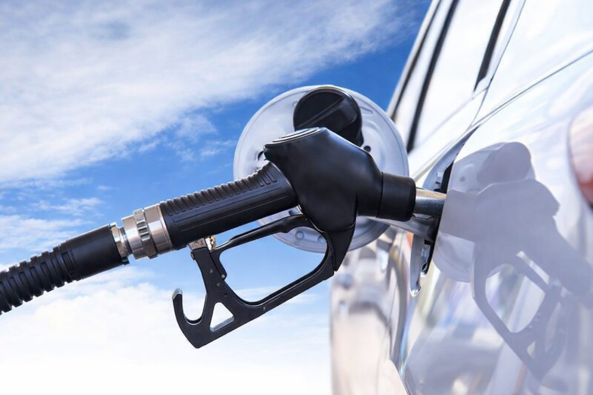 Pistolet de pompe à carburant noir inséré dans l’orifice du réservoir d’une voiture blanche lors d’un plein dans une station-service, vu en gros plan avec le tuyau relié à la pompe et un ciel bleu légèrement nuageux en arrière-plan.