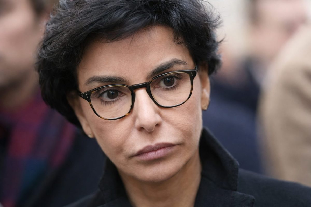 Portrait serré d’une femme politique française portant des lunettes rondes et un manteau sombre, regard sérieux et concentré, photographiée lors d’un événement public avec une foule floue en arrière-plan dans un contexte d’actualité politique et de polémique médiatique.