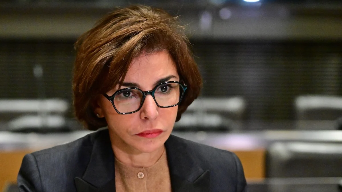 Portrait d’une femme politique au regard fixe et sérieux, portant des lunettes, expression concentrée et déterminée, cadrage serré sur le visage, arrière-plan flou de salle institutionnelle, ambiance tendue évoquant une situation politique importante