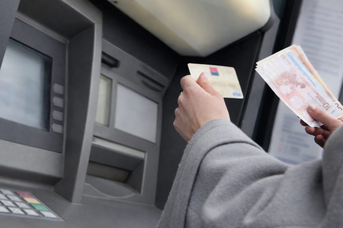 Personne retirant de l’argent à un distributeur automatique de billets, tenant une carte bancaire dans une main et plusieurs billets en euros dans l’autre, devant un guichet automatique dans un espace bancaire.