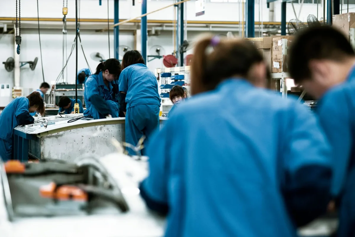 Des ouvriers en tenue de travail bleue s’activent dans un atelier industriel, certains concentrés sur une chaîne de production, illustrant les conditions de travail en usine et les enjeux liés à l’allongement de la durée de travail avec la retraite à 65 ans