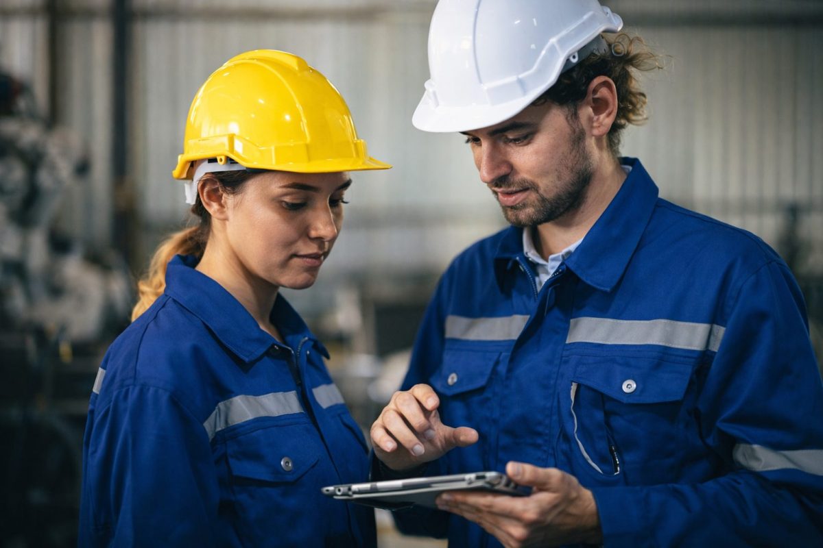 Deux ouvriers en tenue de travail et casques de sécurité consultent une tablette dans un atelier industriel, leurs visages en gros plan illustrant la collaboration et l’allongement de la durée de travail dans le contexte de la retraite à 65 ans