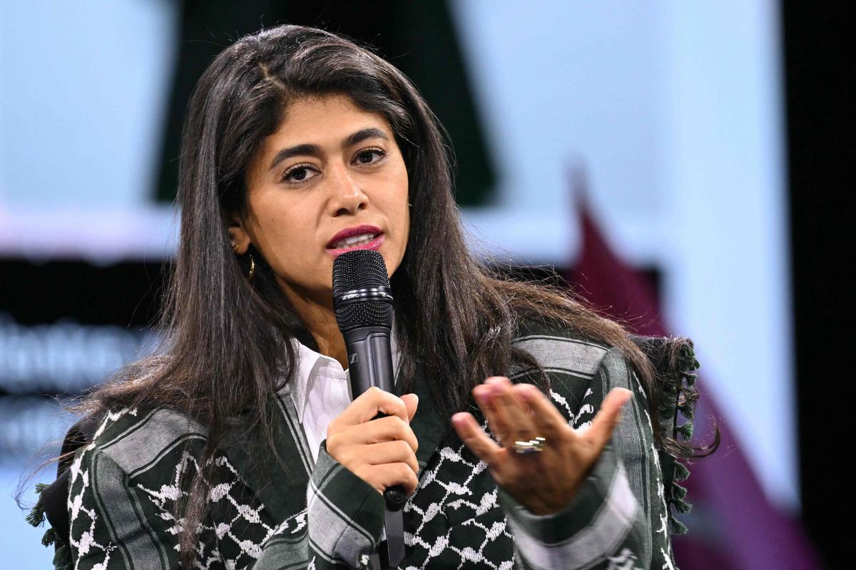 Femme politique s’exprimant avec un micro, regard intense et sérieux, main en mouvement lors d’un discours public, arrière-plan flou de scène, ambiance de tension et de débat