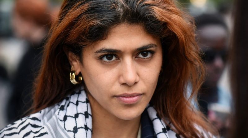 Femme politique au regard intense, visage en gros plan avec expression sérieuse, arrière-plan flou d’une foule en extérieur, ambiance tendue et contexte de controverse politique