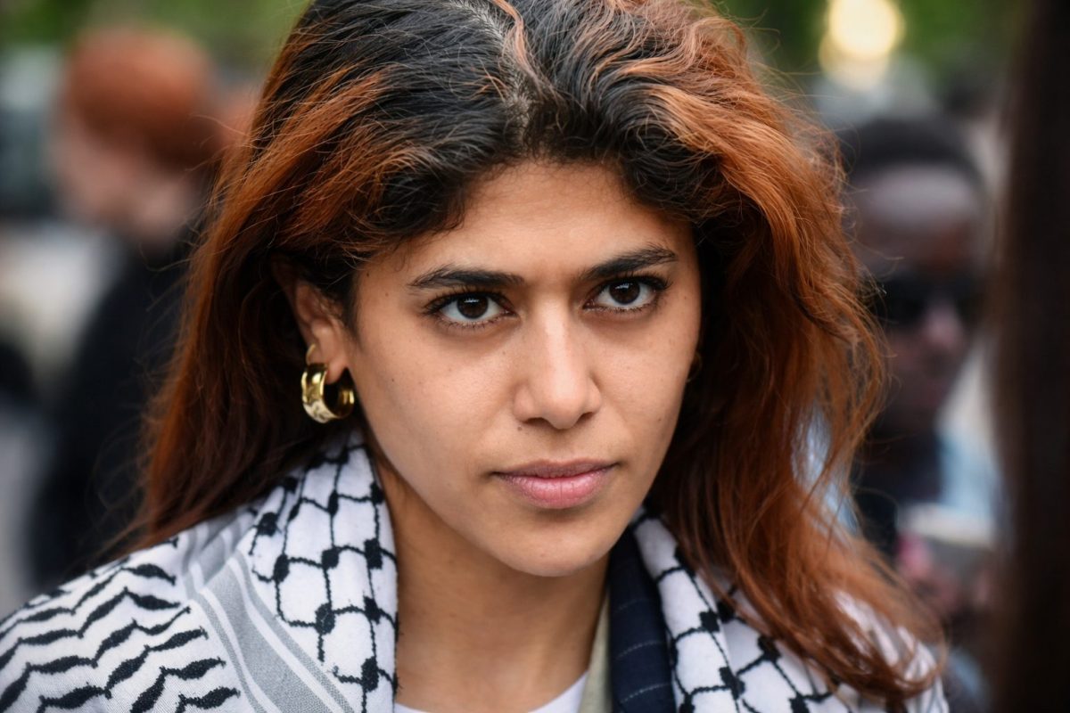 Femme politique au regard intense, visage en gros plan avec expression sérieuse, arrière-plan flou d’une foule en extérieur, ambiance tendue et contexte de controverse politique