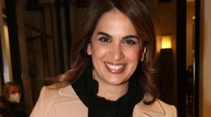 Journaliste souriante en tenue élégante beige et écharpe noire, posant dans un intérieur éclairé aux lumières chaudes, ambiance événement médiatique avec arrière-plan légèrement flou, expression confiante et professionnelle