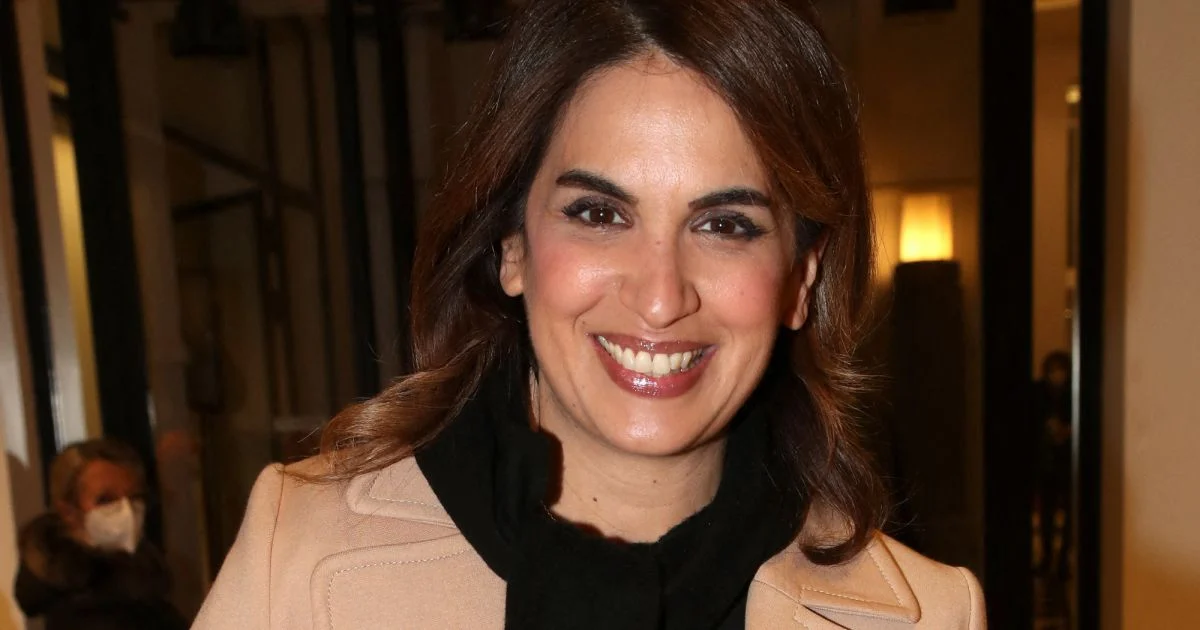 Journaliste souriante en tenue élégante beige et écharpe noire, posant dans un intérieur éclairé aux lumières chaudes, ambiance événement médiatique avec arrière-plan légèrement flou, expression confiante et professionnelle