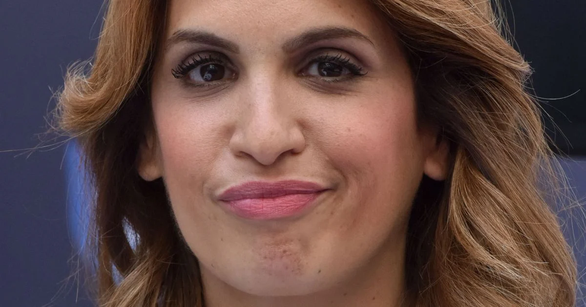 Gros plan sur une journaliste aux cheveux châtains ondulés, maquillage naturel et regard direct face caméra, arrière-plan flou de studio télé aux tons bleutés, expression sérieuse évoquant une prise de parole médiatique
