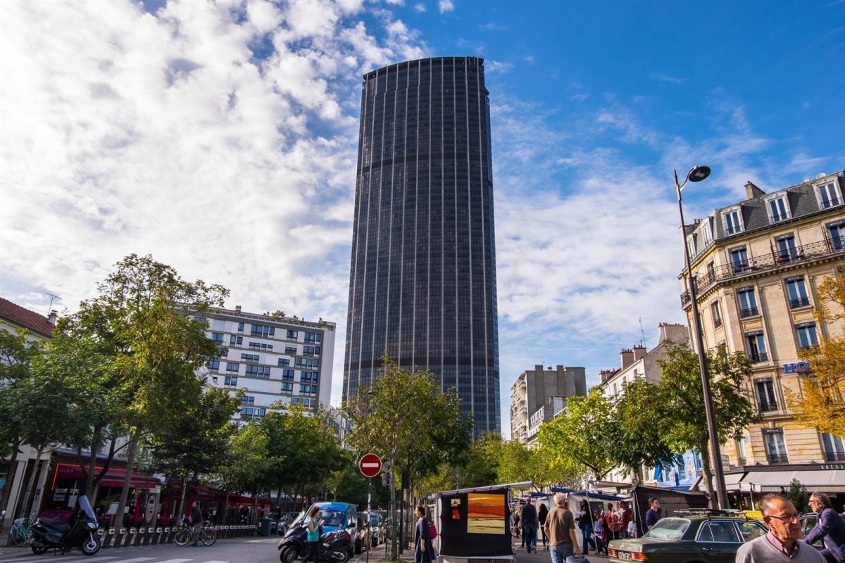 Grand gratte-ciel sombre dominant un quartier urbain avec des immeubles, des arbres et des passants dans la rue sous un ciel bleu partiellement nuageux.