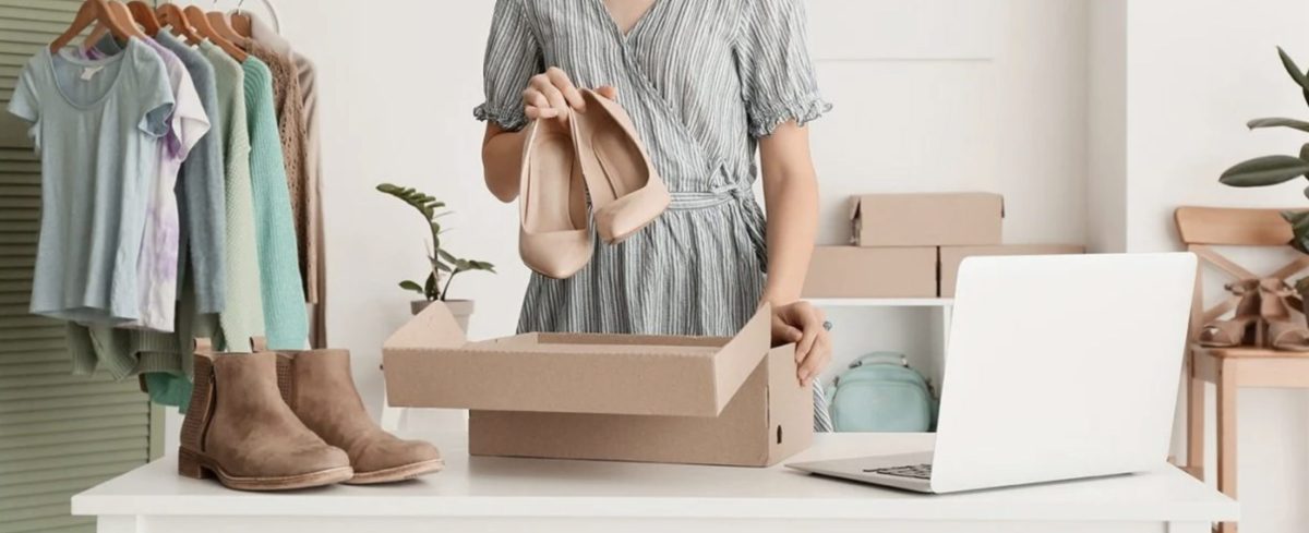 personne préparant l’envoi d’une paire de chaussures d’occasion dans une boîte en carton sur une table, avec un ordinateur portable et des vêtements suspendus sur un portant, illustrant la revente d’objets et vêtements en ligne