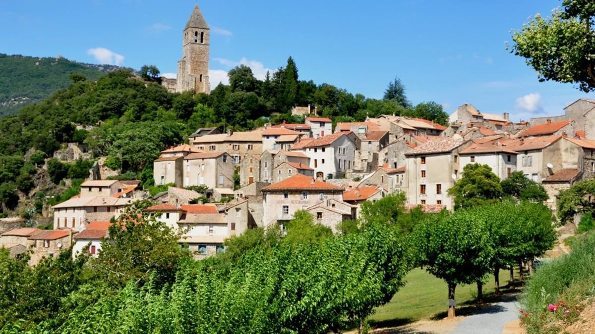 village en pierre construit sur une colline verdoyante avec des maisons aux toits rouges et un clocher dominant le paysage sous un ciel bleu