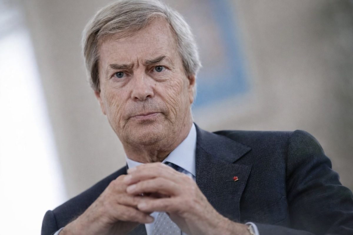 Portrait serré d’un homme d’affaires français au visage sérieux, regard intense et mains jointes, en costume sombre, dans un contexte institutionnel flou évoquant une audition ou une prise de parole officielle