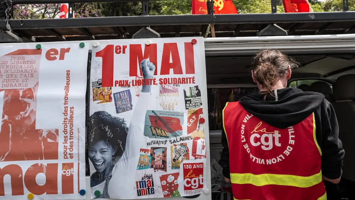Affiches syndicales du 1er Mai collées sur un panneau avec slogans sur les droits des travailleurs et une personne portant un gilet syndical rouge de dos, illustrant une mobilisation sociale