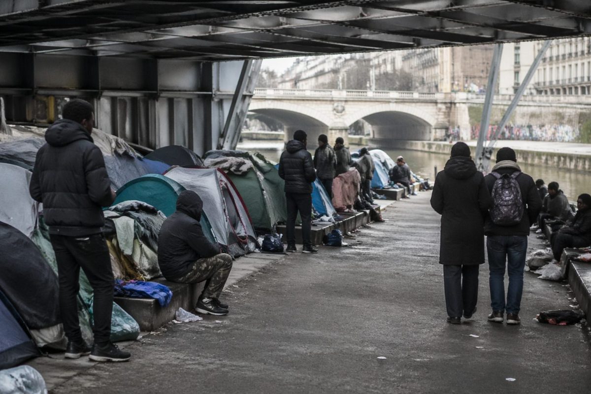 Campement de tentes sous un pont urbain avec plusieurs personnes en situation de précarité, sacs et effets personnels visibles, ambiance grise et tendue illustrant des conditions de vie difficiles en milieu urbain