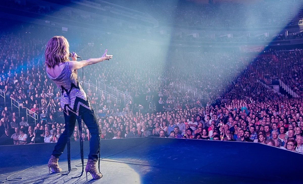 Artiste féminine sur scène vue de dos tenant un micro et tendant le bras vers une immense foule compacte dans une grande salle de concert, éclairages puissants projetant une lumière blanche et bleutée sur le public, spectateurs debout et assis regardant la scène avec attention et enthousiasme, ambiance électrique et immersive avec des milliers de personnes rassemblées pour un spectacle musical très attendu à Paris, illustrant la forte demande et l’engouement autour des concerts populaires et des billets très recherchés