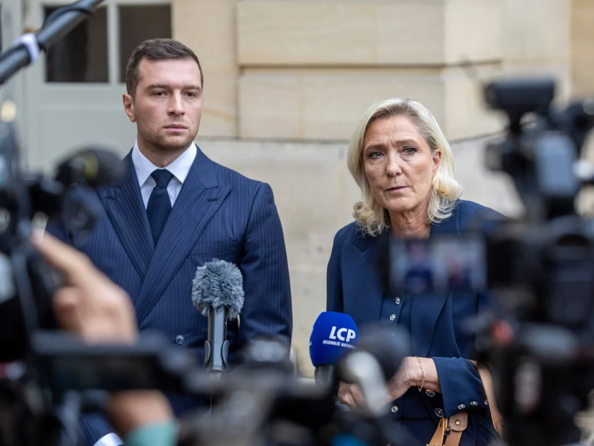 Deux responsables politiques français lors d’une conférence de presse en extérieur, entourés de caméras, micros et journalistes, l’un en costume sombre regardant droit devant, l’autre prenant la parole avec une expression grave, illustrant un contexte de tensions politiques et d’enjeux électoraux à l’approche d’une échéance majeure.