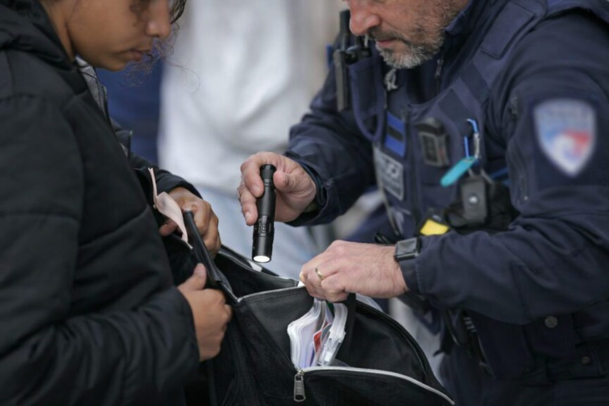Un agent effectue un contrôle minutieux du contenu d’un sac ouvert tenu par une personne, éclairé à l’aide d’une lampe, focus sur les mains et les objets inspectés, ambiance tendue, scène réaliste en extérieur
