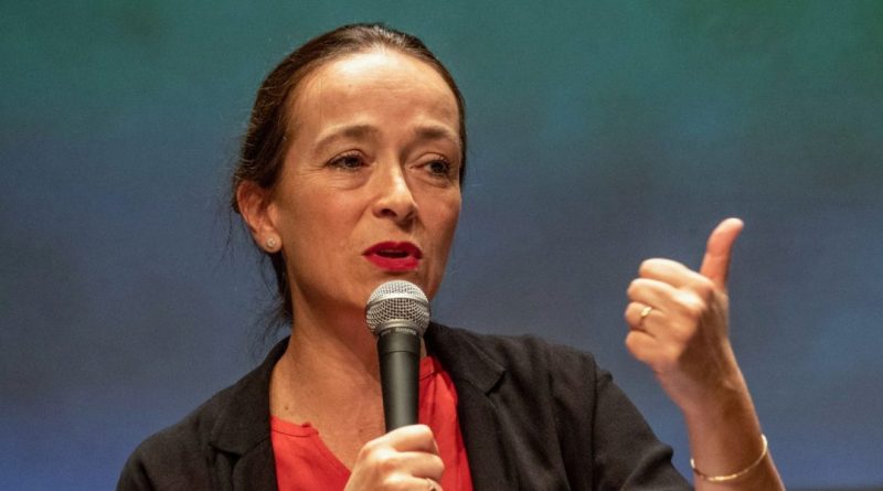 Femme prenant la parole avec un micro, geste de la main affirmé, lors d’une intervention publique dans un contexte professionnel