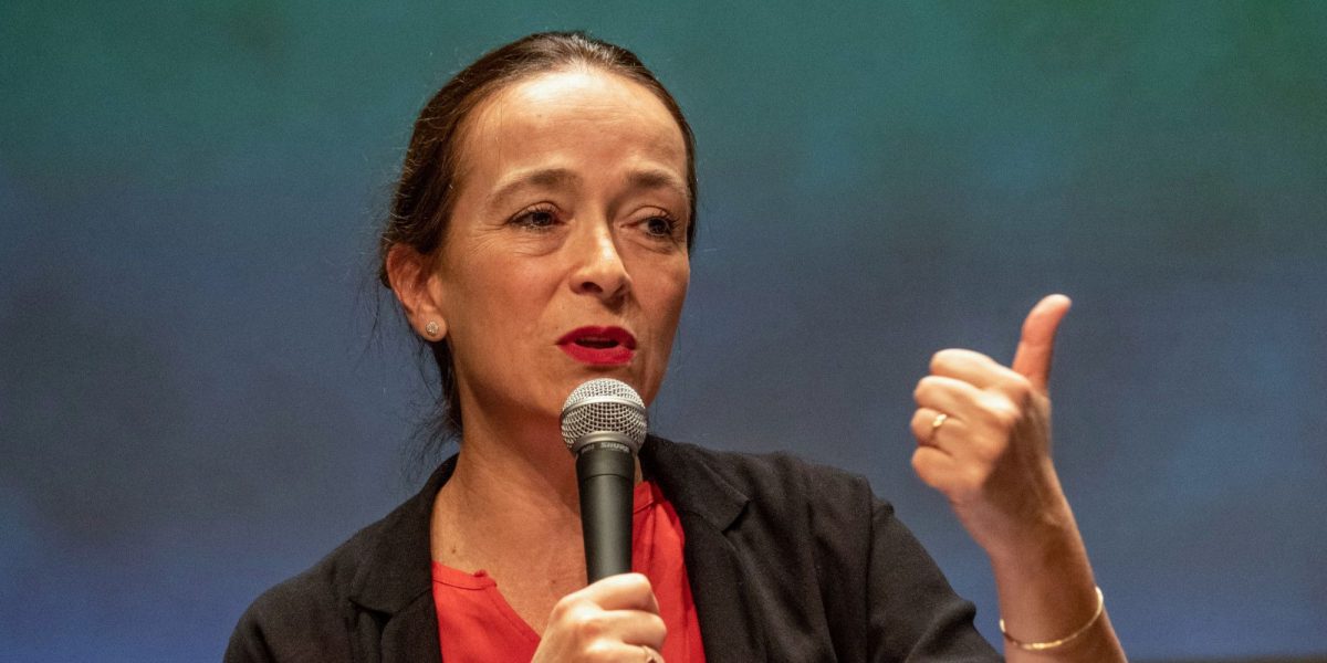 Femme prenant la parole avec un micro, geste de la main affirmé, lors d’une intervention publique dans un contexte professionnel