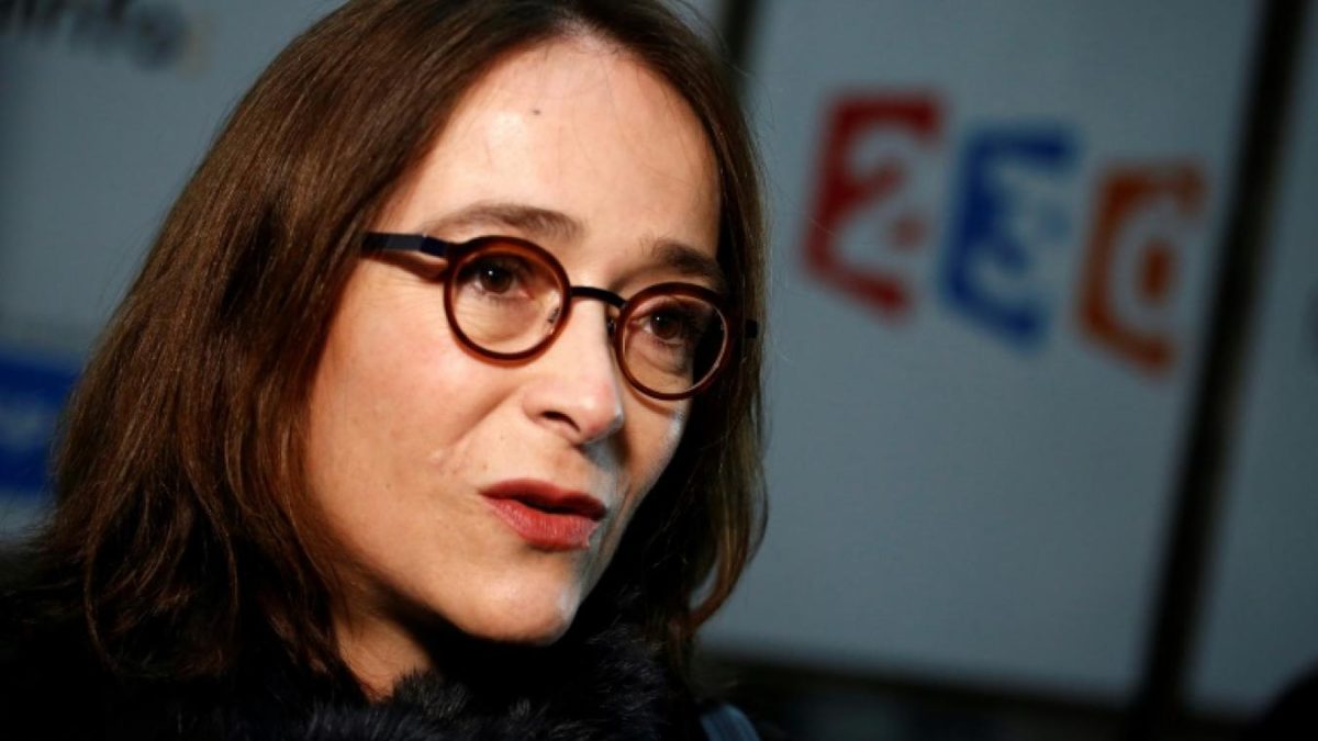 Femme dirigeante du secteur audiovisuel français, en plan serré, portant des lunettes rondes et un manteau sombre, expression sérieuse et concentrée, regard tourné légèrement sur le côté comme lors d’une prise de parole publique, avec en arrière-plan flou un logo de chaîne de télévision aux couleurs bleu, blanc et rouge, illustrant un contexte de polémique médiatique autour de la rémunération et de la gestion du service public audiovisuel en France.