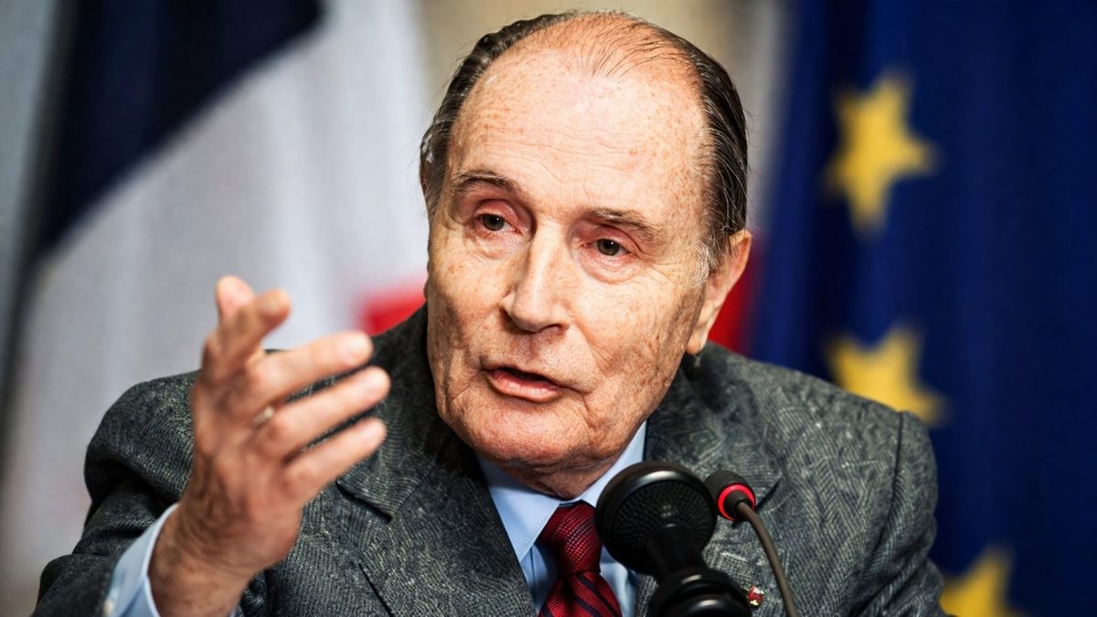 Portrait serré d’un homme politique âgé en costume gris et cravate rouge s’exprimant au micro lors d’une conférence officielle, regard intense et main levée en geste d’insistance, avec des drapeaux flous en arrière-plan