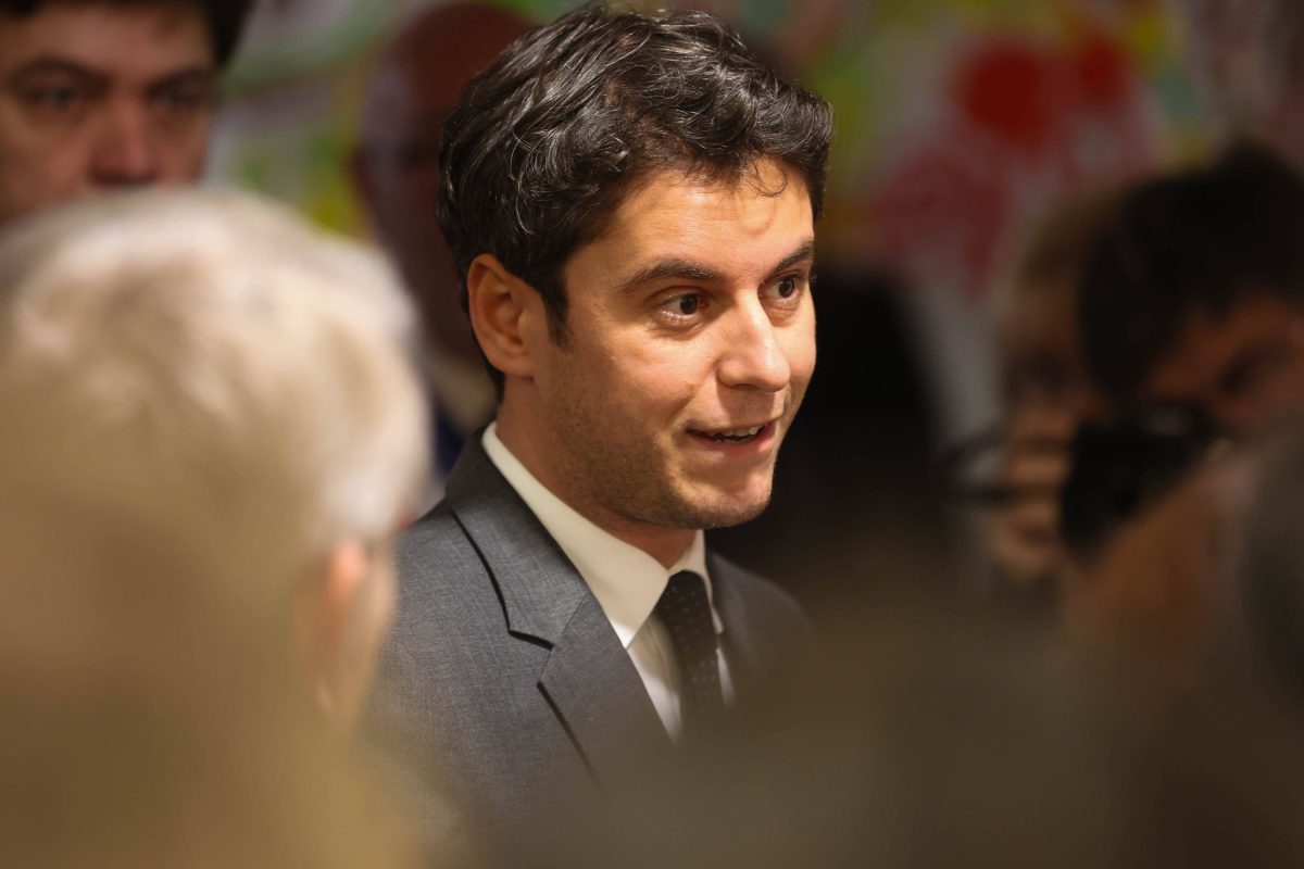 Homme en costume et cravate parlant dans une pièce bondée, regard attentif et expression engagée, entouré de personnes floues, ambiance chaleureuse illustrant une prise de parole publique et un échange direct