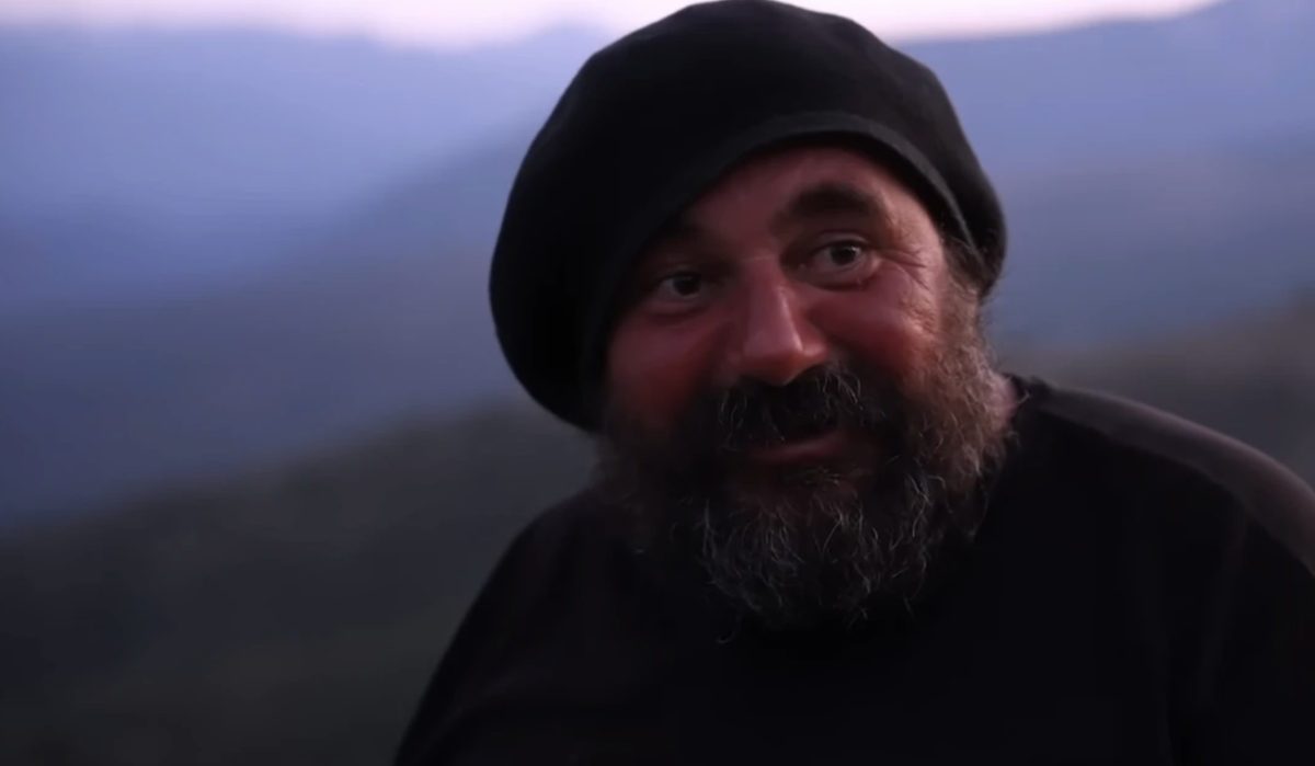 Homme barbu portant un bonnet noir, regard tourné sur le côté, assis en pleine nature dans un environnement montagneux, avec des reliefs flous à l’arrière-plan et une lumière douce de fin de journée, illustrant une vie isolée et paisible loin du monde moderne