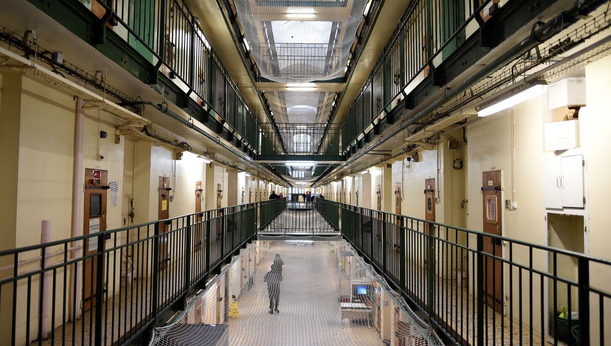 Couloir intérieur d’une prison avec cellules alignées sur plusieurs étages, passerelles métalliques et filet de sécurité au centre