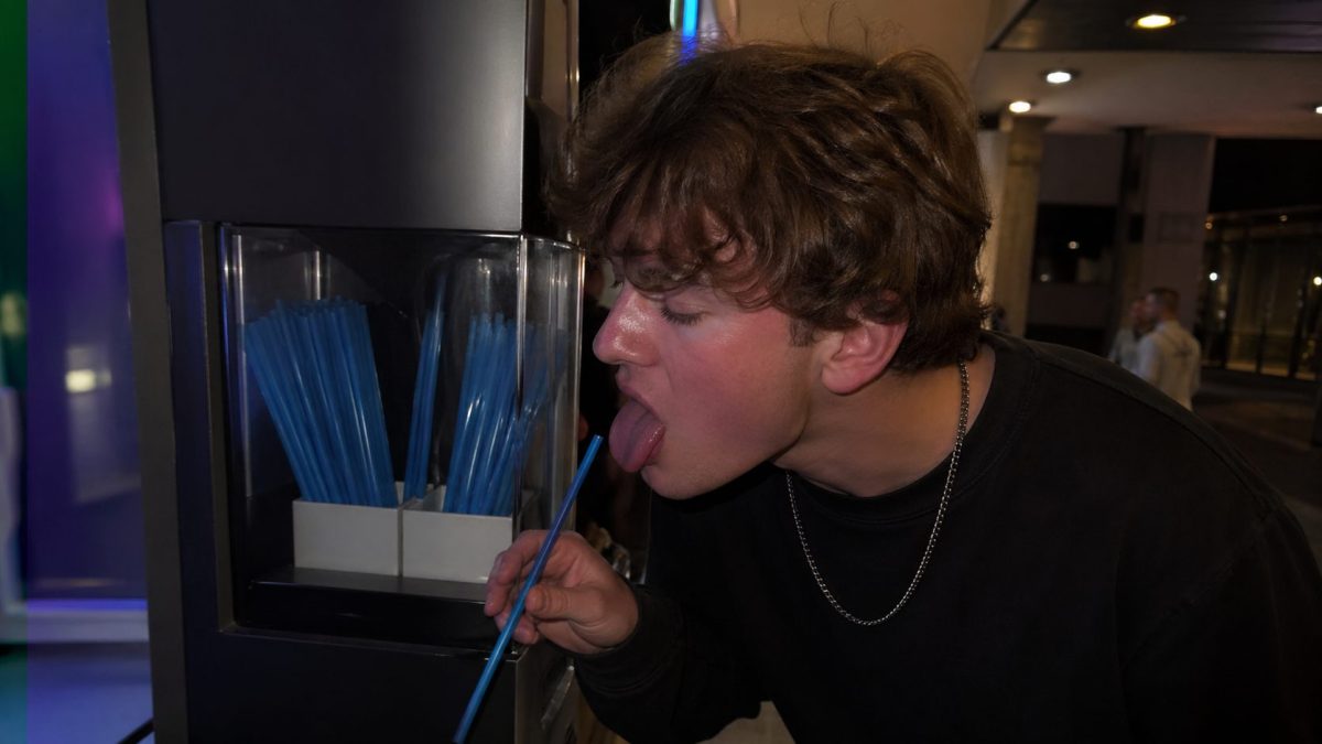 Jeune homme filmant un geste provocant avec une paille dans un distributeur de boissons, scène inquiétante dans un lieu public