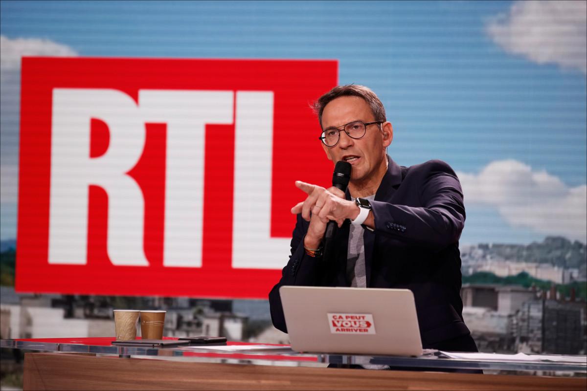 Animateur de radio et télévision tenant un micro sur un plateau avec écran rouge en arrière-plan, en train de s’exprimer avec intensité lors d’une émission en direct