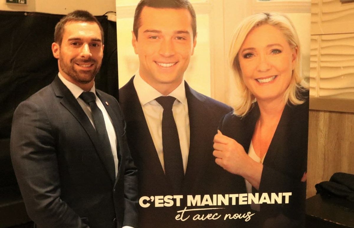 Homme en costume posant à côté d’une grande affiche montrant deux responsables politiques souriants, avec un slogan en arrière-plan, dans un contexte de communication politique