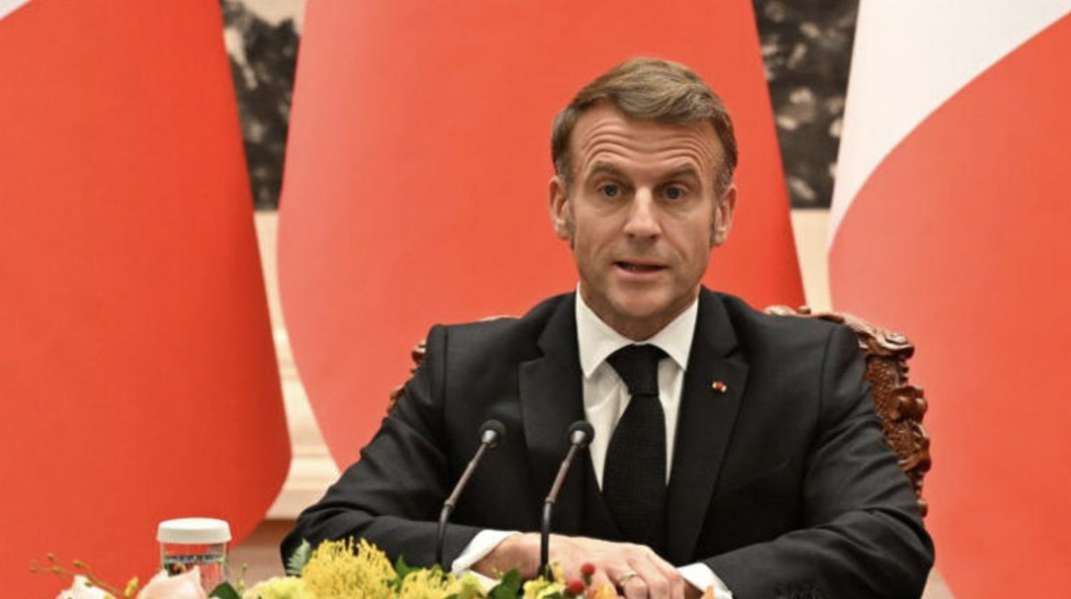 Un homme politique en costume sombre assis derrière un bureau officiel avec deux micros, devant des drapeaux rouges, lors d’une prise de parole solennelle dans un contexte de tension politique en France